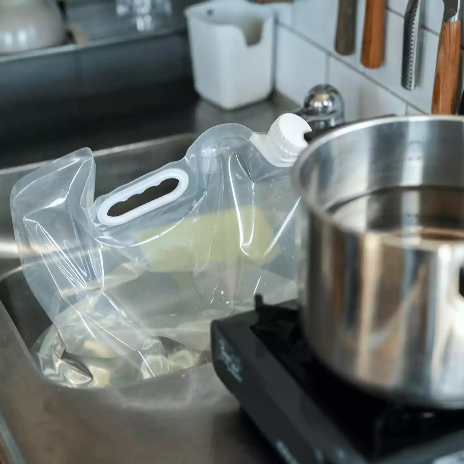 En genomskinlig, hopfällbar vattenbehållare med vit kork och handtag står i en diskho bredvid en kastrull på en kokplatta.