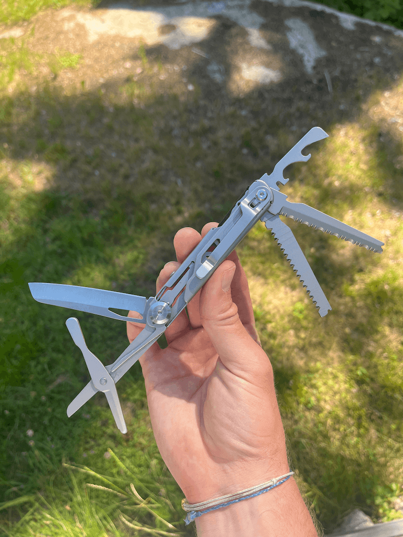 En hand håller upp ett utfällt multiverktyg med kniv, såg och korkskruv, mot en gräsmatta i bakgrunden.