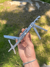 En hand håller upp ett utfällt multiverktyg med kniv, såg och korkskruv, mot en gräsmatta i bakgrunden.
