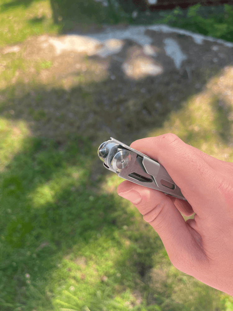 En hand håller ett hopfällt multiverktyg, mot en grönskande gräsmatta med solfläckar.