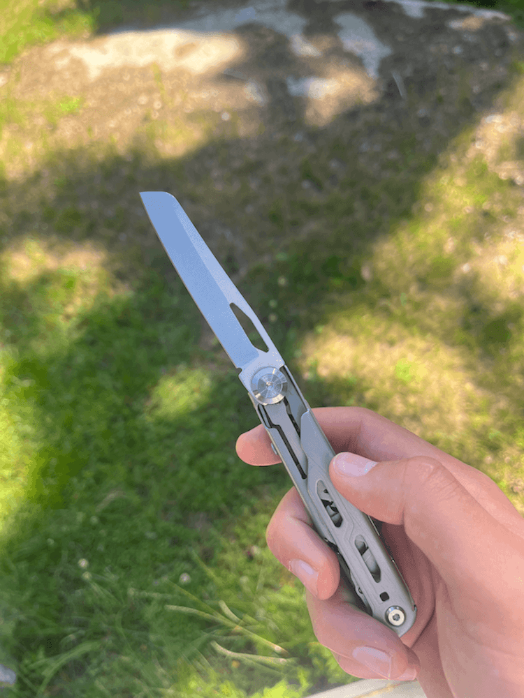 En hand håller ett multiverktyg med en utfälld kniv, mot en solig gräsmatta med skuggor.