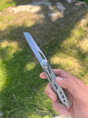 En hand håller ett multiverktyg med en utfälld kniv, mot en solig gräsmatta med skuggor.