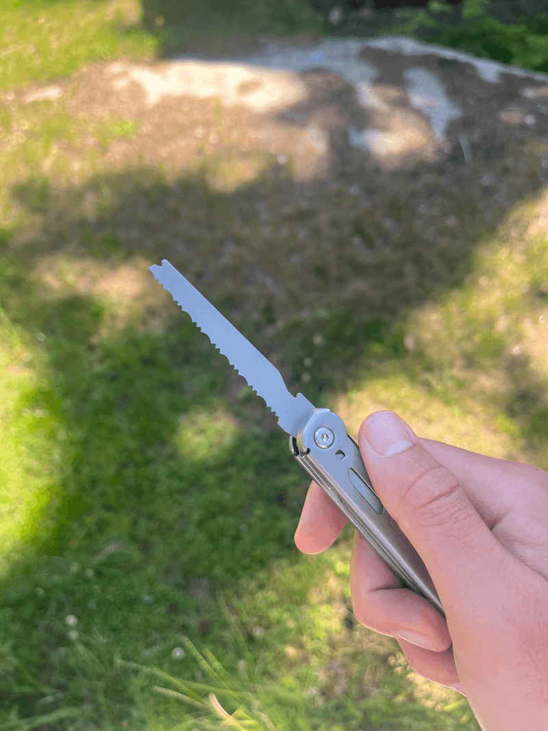 En hand håller ett multiverktyg med en utfälld såg, mot en solig gräsmatta med skuggor.