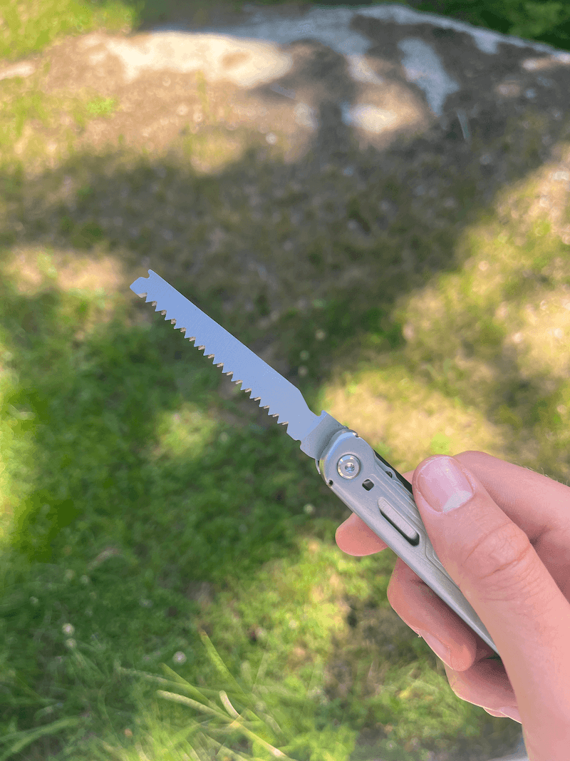 En hand håller ett multiverktyg med utfälld såg, mot en gräsig bakgrund.