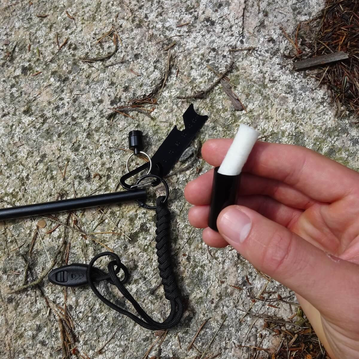 En hand håller ett svart tändstål med vit spets, bredvid en karbinhake och en del av ett verktyg på en stenig mark.