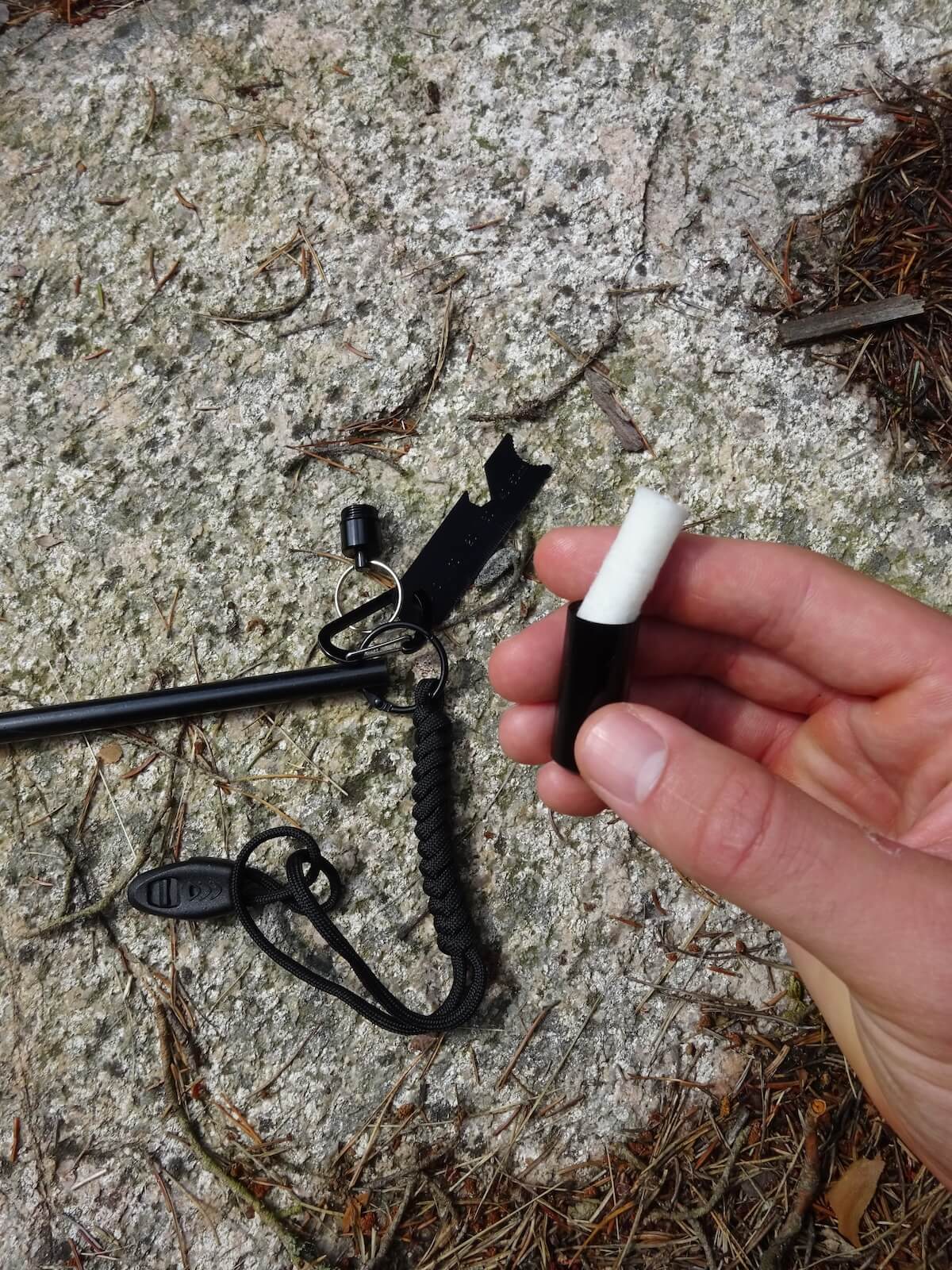 En hand håller ett svart tändstål med vit spets, bredvid en karbinhake och en del av ett verktyg på en stenig mark.